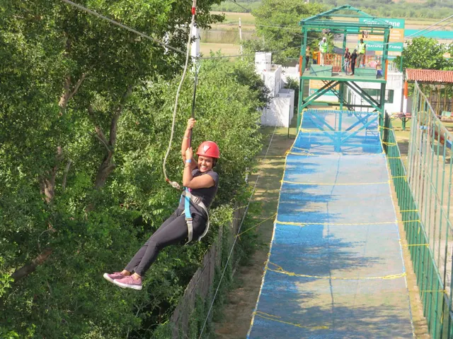 Woman in a red helmet and harness ziplining along a cable above a blue landing mat