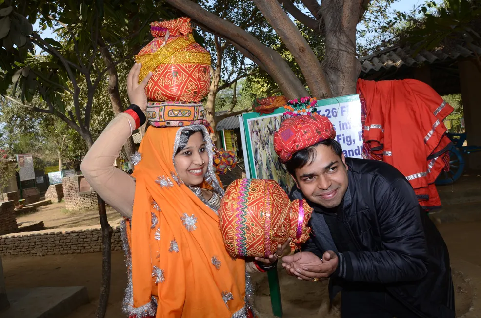 Woman in an orange veil balancing a decorated pot on her head beside a man in a red turban
