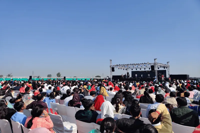 Big outdoor gathering seated in the open facing a lit stage and truss