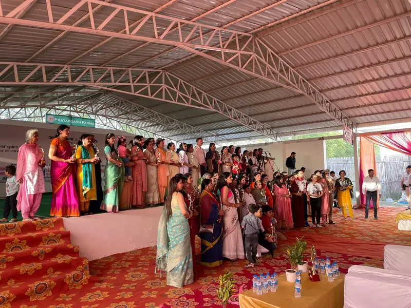 Women in saris holding diyas on stage beneath a Pratapgarh Farms banner