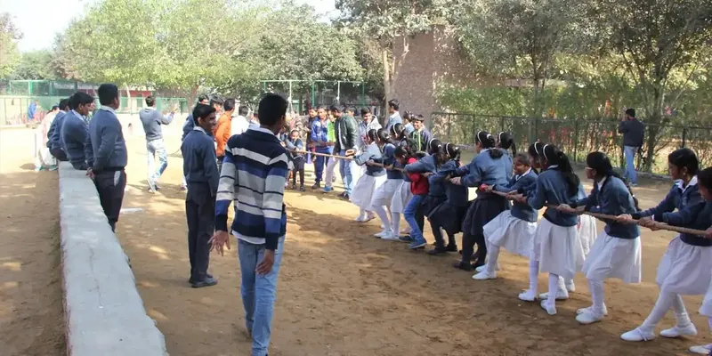 Students in sweaters and white salwar pulling a rope in a tug-of-war on dirt