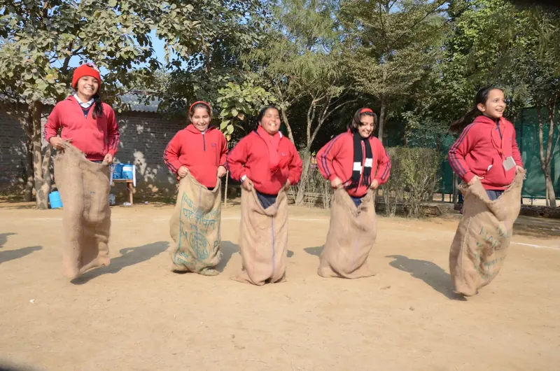 Five girls in red hoodies hopping in a sack race across a dirt ground