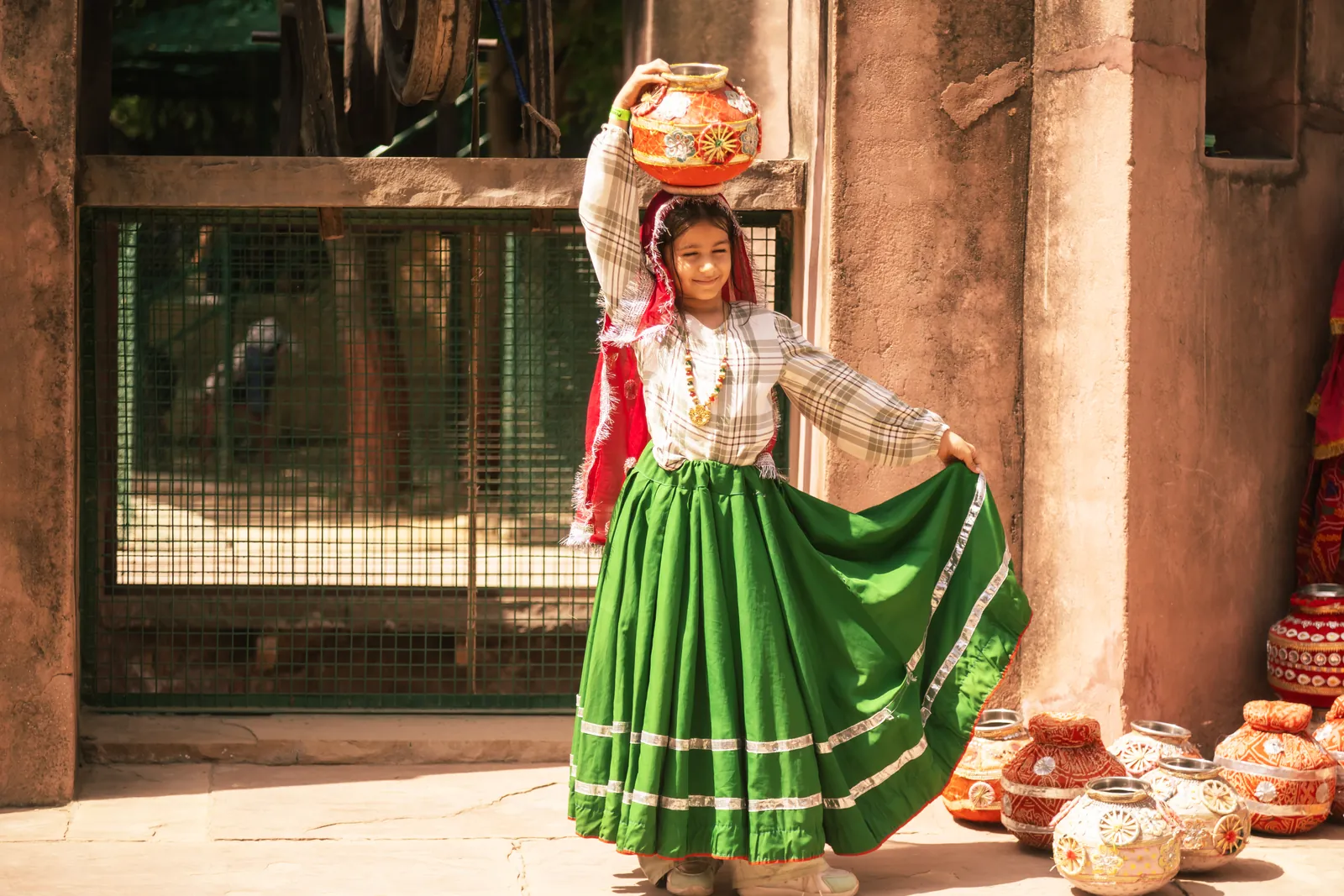 A performer in a green Haryanvi ghaghra balancing stacked pots on her head in the village yard at Pratapgarh Farms, with guests seated on the ground around her