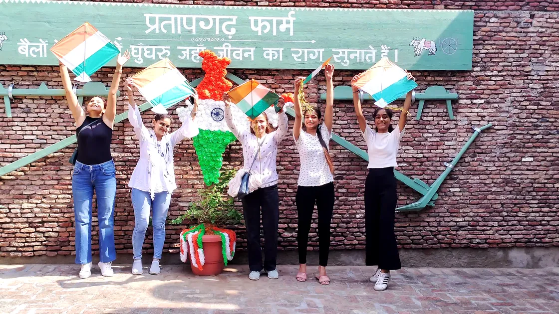 Five women holding kites above their heads before the farm's painted brick wall and tricolour decoration