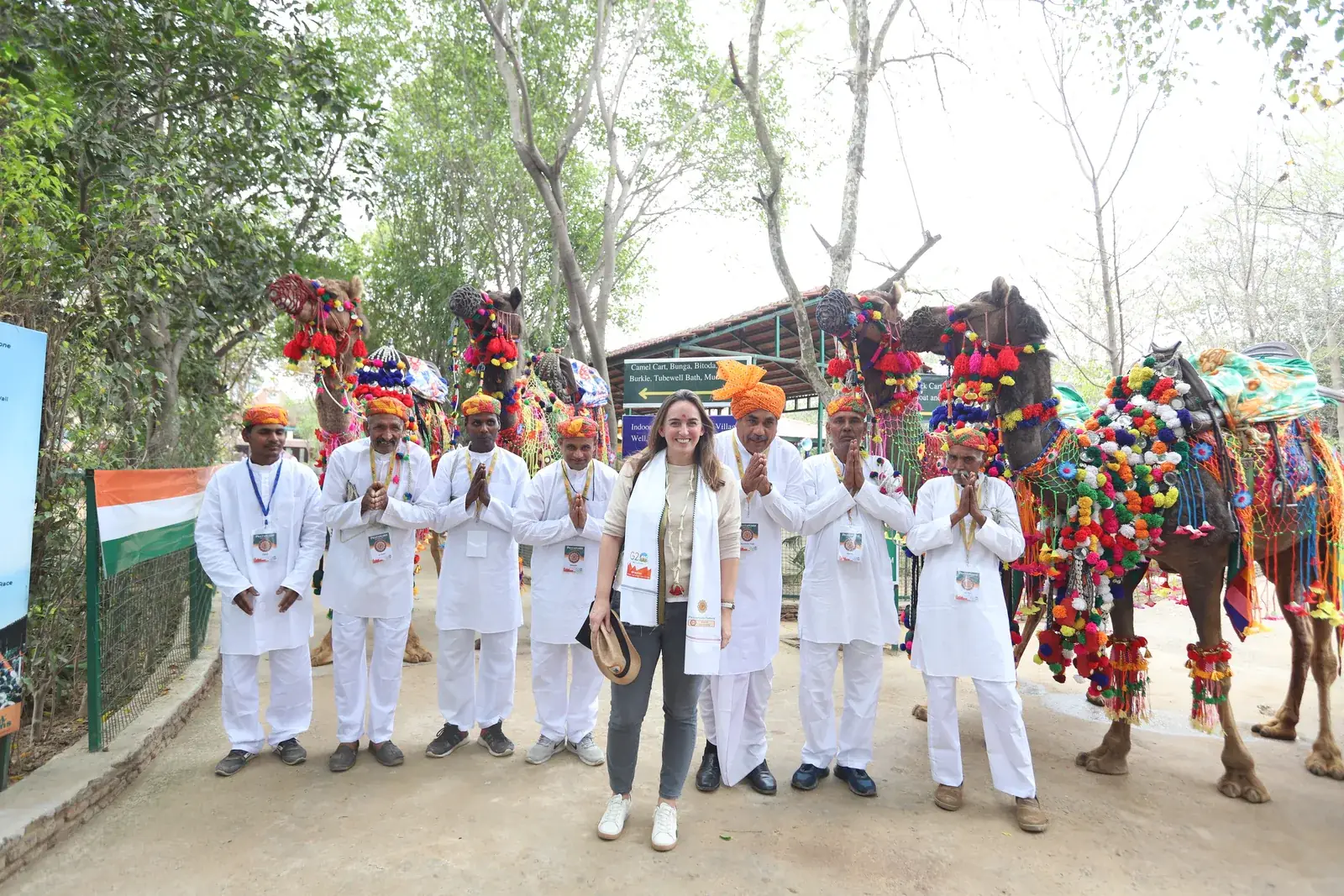 A delegate standing with seven farm hosts in orange pagdis and white kurtas, all with folded hands, in front of three camels decorated with coloured tassels